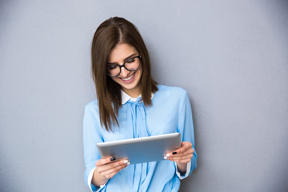 Happy businesswoman standing with table computer over gray background. Wearing in blue shirt and glasses.